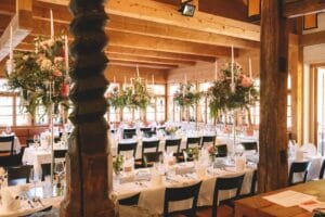 Ein Speisesaal mit Holzbalken und großen Fenstern, perfekt für eine Hochzeit, bietet elegant gedeckte Tische mit weißen Tischdecken und schwarzen Stühlen. Große, üppige Blumengestecke mit Kerzen schmücken die Tische und schaffen eine warme und einladende Atmosphäre.