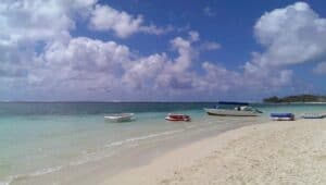 Eine ruhige Strandszene, perfekt für eine traumhafte Hochzeit, mit drei Booten, die an einem sonnigen Tag nah am Ufer vor Anker liegen. Das klare türkisfarbene Wasser trifft sanft auf den Sandstrand unter einem blauen Himmel mit flauschigen weißen Wolken.