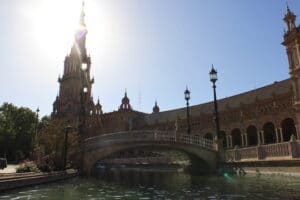 Blick auf die Plaza de España in Sevilla, Spanien, perfekt für eine traumhafte Hochzeit. Das Bild zeigt ein großes, reich verziertes Gebäude mit einem zentralen Turm und einer Steinbrücke über einen Kanal. Die Sonne scheint hell und erzeugt einen Silhouetteneffekt. Bäume und Laternen säumen den Bereich und schaffen eine romantische Kulisse für Hochzeiten.