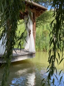Ein hölzerner Pavillon mit durchsichtigen weißen Vorhängen steht über einem ruhigen Teich, umgeben von üppigem Grün und herabhängenden Weidenzweigen. Sonnenlicht fällt hindurch und wirft Reflexionen auf das Wasser – perfekt für alle, die von einer ruhigen Hochzeitskulisse träumen.