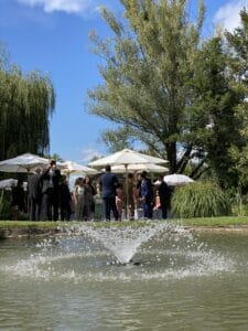 Eine Gruppe formell gekleideter Menschen versammelt sich unter weißen Sonnenschirmen in der Nähe eines Teichs mit einem Springbrunnen und schafft so die perfekte Kulisse für eine Hochzeit in diesem üppigen Garten mit hohen Bäumen und einem klaren blauen Himmel.