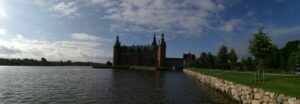 Panoramablick auf Schloss Frederiksborg in Dänemark, dessen markante Türme und Türme ideal für die Hochzeitsplanung sind. Das Schloss, umgeben von einem großen See an einem sonnigen Tag mit vereinzelten Wolken, bietet eine malerische Kulisse. Ein Steinpfad und üppiges Grün rahmen die rechte Seite des Bildes ein.