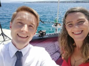 Ein Mann und eine Frau lächeln in die Kamera, möglicherweise während ihrer Hochzeitsplanung. Sie sind draußen in der Nähe eines Gewässers, mit sichtbaren Noten und einer digitalen Tastatur. Der Himmel ist klar und in der Ferne ist Land zu sehen. Beide sind halbformell gekleidet, was auf bevorstehende Hochzeitsfeierlichkeiten hindeutet.