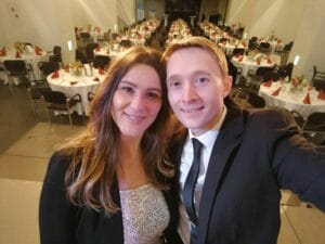 Ein Mann und eine Frau in Abendgarderobe machen ein Selfie bei einem Hochzeitsbankett. Der Saal ist voller runder Tische mit weißen Tischdecken, roten Servietten und Blumendekorationen, die ihre elegante Hochzeitsplanung perfekt widerspiegeln.