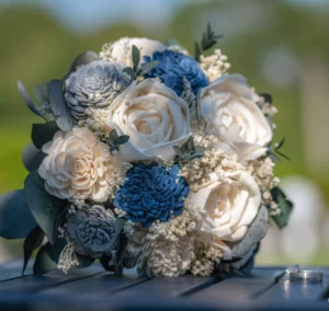Ein Strauß weißer und blauer Blumen mit grünen Blättern auf einer Holzoberfläche weckt Romantik. Der unscharfe Hintergrund lässt an eine Außenkulisse denken, die perfekt für eine Hochzeit ist. Damit ist es die ideale Wahl für alle, die schon mitten in ihrer Hochzeitsplanung stecken.