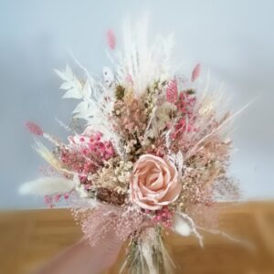 Eine Hand hält einen Strauß Trockenblumen in zarten Rosa-, Weiß- und Beigetönen, perfekt für eine romantische Hochzeit. Das Arrangement besteht aus Rosen, winzigen Knospen, Gräsern und zartem Blattwerk vor einem hellblau-grauen Hintergrund – ideal für Ihre Hochzeitsplanung.