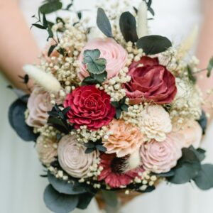 Ein Brautstrauß mit verschiedenen Blumen, darunter rote und rosa Rosen, umgeben von grünen Blättern und kleinen weißen Blüten. Ideal für alle, die gerade mit der Hochzeitsplanung beschäftigt sind. Der Strauß wird von jemandem in einem hellen Kleid gehalten.