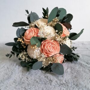 Ein Strauß zartrosa und cremefarbener Rosen ist von Eukalyptusblättern und kleinen weißen Blüten umgeben, die hübsch auf einer weichen, hellgrauen Stoffoberfläche arrangiert sind – perfekt für eine verträumte Hochzeitsfeier.
