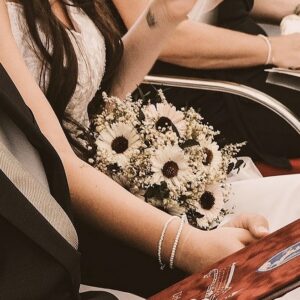 Eine Person, die einen Strauß weißer Gänseblümchen und Grünzeug hält, sitzt auf roten Stühlen und strahlt in einem weißen Kleid mit langen dunklen Haaren Eleganz aus. Es ist eine malerische Hochzeitskulisse, mit anderen Gästen, die sich im Hintergrund unterhalten.