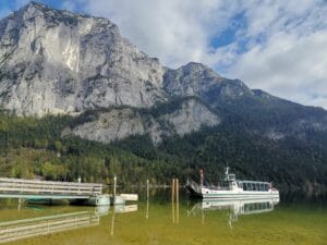 Ein malerischer Blick auf ein Boot, das an einem klaren, ruhigen See angedockt ist, umgeben von hohen, schroffen Bergen und einem Wald. Perfekt für diejenigen, die inmitten der Schönheit der Natur heiraten möchten. Der Himmel ist teilweise bewölkt, was dieser malerischen Landschaft Tiefe verleiht.