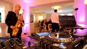 Eine lebendige Hochzeitsfeierszene zeigt im Vordergrund ein DJ-Setup mit Plattenspielern und Tonmischpulten. Im Hintergrund spielt ein Saxophonist, während die Gäste unter warmem rosa und gelbem Licht plaudern, was sich nahtlos in die sorgfältige Hochzeitsplanung einfügt.