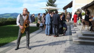 Ein Saxophonist spielt im Freien bei einer Hochzeitsfeier. Mehrere Personen in formeller Kleidung stehen in Gruppen in der Nähe eines Gebäudes mit einer malerischen Kulisse aus Hügeln und Bäumen. Auf der Terrasse sind Tische mit Sonnenschirmen zu sehen, die der Hochzeitsplanungsveranstaltung Charme verleihen.