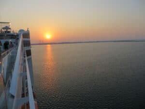 Vom Deck des Kreuzfahrtschiffs aus erstreckt sich bei Sonnenuntergang ein ruhiges Meer unter einer goldenen Spiegelung. Während die Sonne am Horizont versinkt, besprechen Paare verträumt ihre Hochzeitspläne und genießen dabei die romantische Aussicht.