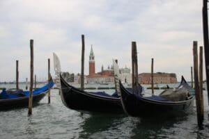 Am Canale Grande in Venedig, Italien, angedockte Gondeln bieten mit dem hohen Glockenturm und den historischen Gebäuden der Insel San Giorgio Maggiore im Hintergrund unter einem bewölkten Himmel eine romantische Kulisse, die sich perfekt für Hochzeitsfotos eignet.