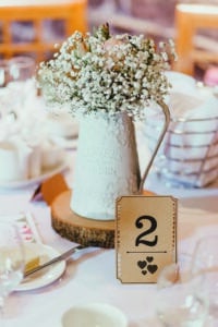 Ein rustikales Tischdekor für eine Hochzeit besteht aus einer weißen Vase mit dem Text „Flowers & Garden“, in der weiße Schleierkraut- und Pfirsichrosen blühen. Es steht elegant auf einer Holzplatte neben einer Tischnummernkarte mit der Zahl 2 und Herzsymbolen.
