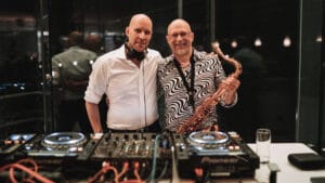 Zwei kahlköpfige Männer stehen lächelnd hinter dem DJ-Equipment bei einer Hochzeit. Einer trägt ein weißes Hemd und hat Kopfhörer um den Hals, der andere ein gemustertes Hemd und hält ein Saxophon. Sie befinden sich in einem schwach beleuchteten Raum mit sichtbaren Spiegelungen im Hintergrund, die ein unvergessliches Ereignis perfekt einfangen.