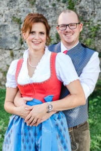 Ein lächelndes Paar, die Frau trägt ein traditionelles rot-blaues Dirndl und der Mann ein weißes Hemd und eine graue Weste. Sie umarmen sich draußen vor einer Steinmauer im Hintergrund und verkörpern die Freude der Hochzeitsplanung, während das Gras zu ihren Füßen sichtbar ist.
