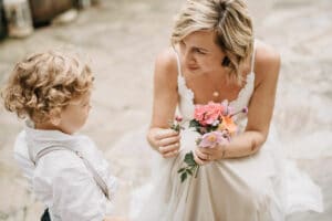 Eine Frau im weißen Kleid lächelt, während sie einem kleinen Jungen im weißen Hemd und Hosenträgern einen kleinen Strauß bunter Blumen zeigt. Ihr freudiger Moment auf dem Steinweg fängt die Schönheit von Hochzeit ein, wo die Liebe unter freiem Himmel erblüht.