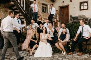 Es spielt sich eine fröhliche Hochzeitsszene ab, mit einer Braut in einem weißen Kleid und ihrem Bräutigam, umgeben von Gästen. Auf rustikalen Steinstufen erfüllen Gelächter und Geplauder die Luft, während sie ihre Hochzeit im Freien feiern, mit einem charmanten Gebäude im Hintergrund.