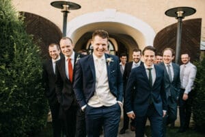 Eine Gruppe von Männern in Anzügen, die lächelnd auf die Kamera zulaufen, strahlen Aufregung aus, während sie an einer Hochzeit teilnehmen. Der Mann in der Mitte trägt einen blauen Anzug und eine weiße Weste; die anderen tragen verschiedene Anzüge und Krawatten. Sie stehen vor einem Gebäude mit Holztüren und Grünflächen auf beiden Seiten.