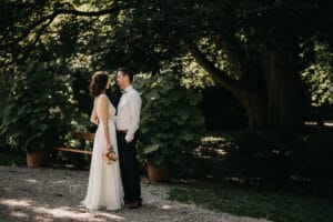 Ein Paar steht sich im Schatten der Bäume in einem Garten gegenüber, eine perfekte Kulisse für ihre Hochzeit. Die Frau trägt ein fließendes weißes Kleid und hält einen Blumenstrauß, während der Mann ein weißes Hemd und dunkle Hosen trägt. Üppiges Grün umgibt sie und schafft eine bezaubernde Kulisse für ihre Liebe.