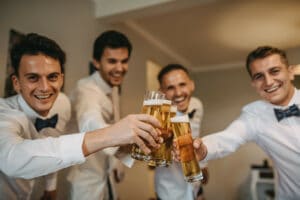 Vier Männer in weißen Hemden und Fliegen lächeln und stoßen mit Biergläsern auf einen freudigen Anlass an. Es sieht so aus, als würden sie die fröhliche Atmosphäre im Haus genießen, vielleicht als Teil der Hochzeitsfeierlichkeiten oder Heiratsvorbereitungen. Ein Hoch auf unvergessliche gemeinsame Momente!