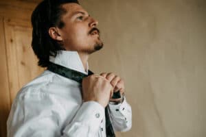 Ein Mann mit langen Haaren und Schnurrbart richtet seine Krawatte, vielleicht bereitet er sich auf ein bedeutendes Ereignis wie eine Hochzeit vor. Er trägt ein weißes Oberhemd mit Manschettenknöpfen und eine dekorative Krawatte. Der neutrale Hintergrund mit einer Holztür zu seiner Linken bereitet die Bühne für den entscheidenden Moment seiner Hochzeitsplanung.