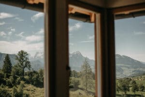 Durch das Fenster entfaltet sich eine malerische Berglandschaft mit üppig grünen Bäumen und Hügeln unter einem wolkig blauen Himmel – eine ideale Kulisse für alle, die davon träumen, inmitten der Pracht der Natur zu heiraten.