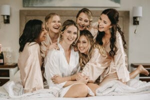 Sechs Frauen in Seidenroben sitzen und lachen zusammen auf einem Bett, fröhlich und entspannt in ihrer gemütlichen Umgebung. Inmitten des Gelächters liegt ein Hauch von Heiraten in der Luft, und auf dem Nachttisch steht eine Flasche Champagner für die Hochzeitsfeier bereit.