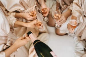 Eine Gruppe von Menschen in Satinroben erhebt Champagnergläser mit Roséwein, während eine andere Person aus einer Flasche einschenkt. Die Kulisse strahlt die Freude der Hochzeitsplanung aus und fängt die Essenz eines herzlichen Zusammenseins in Erwartung einer bezaubernden Hochzeitsfeier ein.