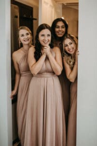 Vier Frauen in passenden beigen Kleidern stehen dicht beieinander in einem Türrahmen, lächeln und blicken in eine Richtung. Die fröhliche Atmosphäre lässt vermuten, dass sie eine Hochzeit feiern und einen besonderen Moment in der Inneneinrichtung festhalten.