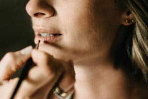 Eine Nahaufnahme einer Person, die mit einem Pinsel Lippenstift aufträgt und so ihren Look für die Hochzeit perfektioniert. Der Fokus liegt auf ihren Lippen und ihrer Hand, die den Pinsel vorsichtig hält. Die Person hat kurzes, helles Haar und trägt einen kleinen Ohrstecker, bereit für den großen Tag voller Liebe und Planung.