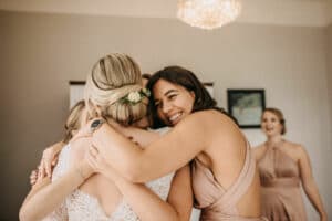 Eine freudige Gruppenumarmung bei einer Hochzeit. Eine Frau im Brautjungfernkleid lächelt herzlich und umarmt die Braut in einem Spitzenkleid mit einem Haarschmuck mit Blumenmotiv. Im Hintergrund sind zwei weitere Brautjungfern zu sehen, ebenfalls in pastellfarbenen Kleidern, die den Moment miterleben.