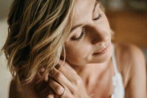 Eine Frau mit kurzen, blonden Haaren und geschlossenen Augen berührt sanft ihren Ohrring und präsentiert einen Verlobungsring an ihrer Hand. Das Bild fängt einen ruhigen, intimen Moment ein, der an eine freudige Hochzeit erinnert, mit sanfter Beleuchtung und einem verschwommenen Hintergrund.