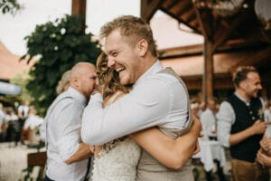 Ein lächelnder Mann in einer hellgrauen Weste umarmt eine Frau in einem weißen Kleid bei einer Hochzeit im Freien. Im Hintergrund sind weitere Gäste in legerer Kleidung zu sehen, die die fröhliche und festliche Atmosphäre dieses besonderen Anlasses unterstreichen.