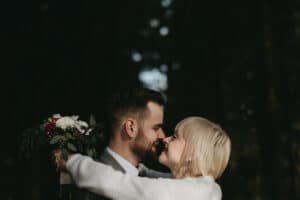 Ein Paar umarmt sich in einem Wald, wobei die Frau einen Strauß weißer und rosa Blumen hält. Die Szene ist warm beleuchtet und fängt einen Moment der Nähe ein, als sie sich vorbeugen, um ihre Nasen zu berühren, und beide sanft lächeln – ein intimer Einblick in ihre Hochzeitsreise.
