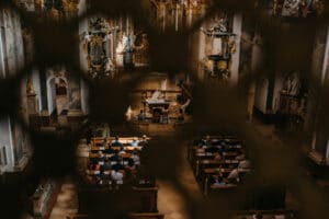 Blick durch das Gitter eines barocken Kircheninneren, reich verziert mit Dekorationen und Statuen. Eine Gemeinde sitzt in den Bänken gegenüber dem Altar, wo Geistliche einen feierlichen Gottesdienst abhalten. Die gedämpfte Beleuchtung trägt zur Atmosphäre bei und schafft eine malerische Kulisse für diejenigen, die hier ihre Hochzeit planen.