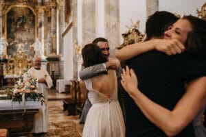 In einer kunstvoll verzierten Kirche mit goldenen Verzierungen umarmt sich ein Paar freudig ein anderes Paar und feiert seine Hochzeit. Ein Priester steht in der Nähe und lächelt neben einem Blumenarrangement auf einem Podest. Die Atmosphäre ist warm und erfüllt von der Aufregung der Hochzeit.