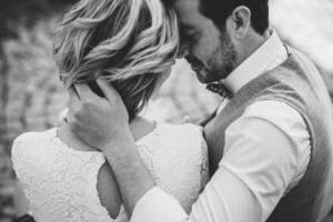 Ein Schwarzweißfoto zeigt ein Paar, das sich eng umarmt und die Essenz einer Hochzeit verkörpert. Der Mann in Oberhemd und Weste hält sanft den Kopf der Frau, die ein Spitzenkleid trägt. Sie teilen einen intimen Moment, der an eine sorgfältige Hochzeitsplanung erinnert, während ihre Umgebung verschwommen ist.