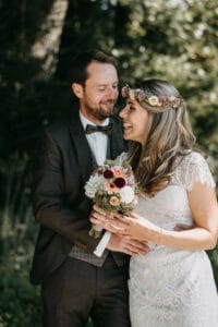 Das Paar steht draußen und lächelt sich an seinem Hochzeitstag an. Die Frau in einem weißen Spitzenkleid und einem Blumenkopfschmuck hält einen Blumenstrauß. Der Mann, der einen dunklen Anzug und eine Fliege trägt, umarmt sie sanft, während Sonnenlicht durch die Bäume hinter ihnen fällt.