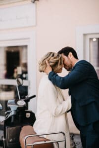 Ein Paar umarmt sich liebevoll vor einem Oldtimer-Roller, der vor einem Gebäude geparkt ist, und fängt einen intimen Moment ihrer Hochzeit ein. Der Mann hält sanft den Kopf der Frau, beide sind stilvoll gekleidet – die Frau in Weiß und der Mann in Dunkelblau – und verkörpern perfekt die Freude des Heiratens.