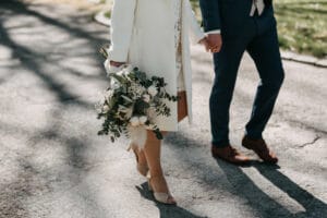 Ein Paar spaziert Hand in Hand an einem sonnigen Tag, vielleicht inmitten ihrer Hochzeitsplanung. Die Frau, in einen weißen Mantel gehüllt, trägt einen Strauß weißer und grüner Blumen. Der Mann, in einem blauen Anzug, steht ihrer Eleganz in nichts nach. Auf dem sonnenbeschienenen Weg sind nur ihre Unterkörper zu sehen.