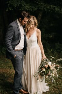 In einer ruhigen Waldgegend bereiten sich Braut und Bräutigam auf ihre Hochzeit vor. Die Braut in einem fließenden weißen Kleid umarmt die Natur mit ihrem Blumenstrauß und Grünpflanzen. Der Bräutigam ergänzt sie in einem grauen Anzug mit braunen Schuhen. Gemeinsam blicken sie nach unten und strahlen Ruhe und Glück aus.