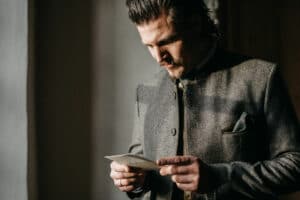 Ein Mann in einer grauen Jacke steht drinnen und betrachtet aufmerksam ein Stück Papier zur Hochzeitsplanung. Das Licht wirft Schatten und hebt seinen konzentrierten Gesichtsausdruck hervor, während er sich auf die bevorstehende Hochzeit vorbereitet.