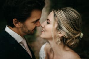 Während der Hochzeitsplanung genießt ein Paar in formeller Kleidung einen intimen Moment im Freien. Die Frau, deren helles Haar hochgesteckt und mit aufwendigen Ohrringen geschmückt ist, lächelt sanft, während sie sich zueinander neigen. Der Mann hat dunkles Haar und trägt Anzug und Krawatte.