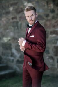 Ein Mann in einem burgunderfarbenen Smoking mit weißem Einstecktuch und schwarzer Fliege steht draußen und richtet sein Jackett und verkörpert damit die Essenz des Heiratens. Im Hintergrund ist eine verschwommene Steinmauer zu sehen, die zur formellen und eleganten Hochzeitsatmosphäre beiträgt.