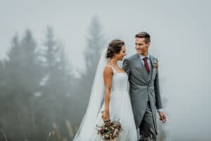 Ein Paar im Hochzeitskleid steht an einem nebligen Tag im Freien und verkörpert die Freude des Heiratens. Die Braut in einem weißen Kleid mit einem Blumenstrauß in der Hand lächelt ihren Bräutigam in einem grauen Anzug mit roter Krawatte an. Sie tauschen einen liebevollen Blick inmitten verschwommener Bäume im Hintergrund – ein perfekter Schnappschuss für ihr Hochzeitsalbum.