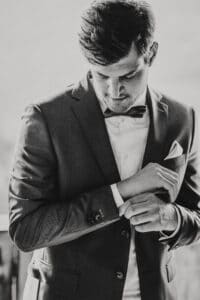 Ein Mann im Anzug rückt seine Manschettenknöpfe zurecht und bereitet sich auf einen eleganten Moment bei seiner Hochzeit vor. Er trägt eine Fliege und hat ein Einstecktuch in seinem Jackett. Das zeitlose Schwarzweißbild fängt die Essenz formeller Eleganz ein und passt perfekt zu einem so besonderen Anlass.