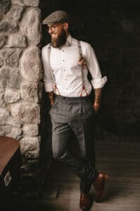 Ein Mann mit Bart lehnt an einer Steinmauer und verkörpert den Geist einer Vintage-Hochzeit. Er trägt eine Schiebermütze, eine Brille, ein weißes Hemd mit Hosenträgern, karierte Hosen und braune Schuhe. Ein kleiner Knopfverschluss ziert sein Hemd und er wirkt entspannt und lächelnd.