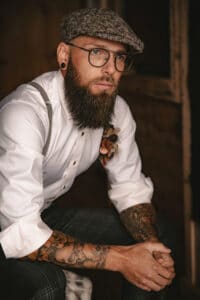 Ein bärtiger Mann mit Brille und Tattoos sitzt nachdenklich da und verkörpert die Essenz des Hochzeitsstils. Er trägt eine Schiebermütze, ein weißes Hemd mit Hosenträgern und dunkle Hosen. An seinem Hemd ist eine Blumenboutonniere befestigt. Der schwach beleuchtete Hintergrund verleiht eine rustikale Atmosphäre, die zu jeder Hochzeitsplanung passt.
